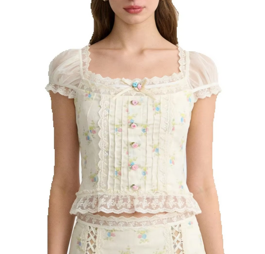 A model wearing a white lace-trimmed blouse with a delicate pastel floral print, featuring a square neckline, sheer puff sleeves, and decorative buttons down the front.