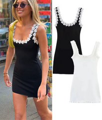 A woman wearing a black mini dress with white floral appliques on the shoulder straps, holding a yellow bag.