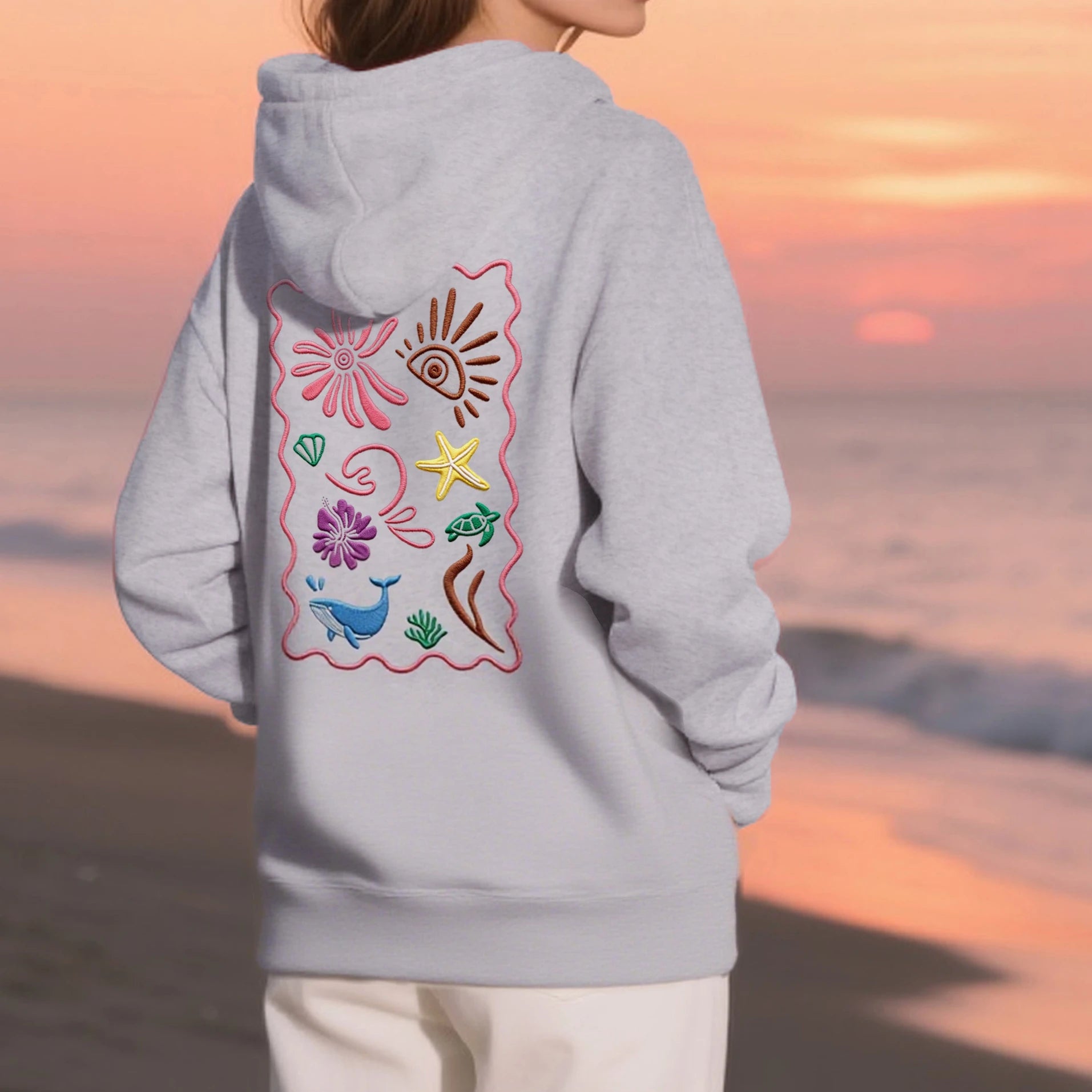 Person wearing a gray hoodie with colorful designs on the back, standing on a beach at sunset.