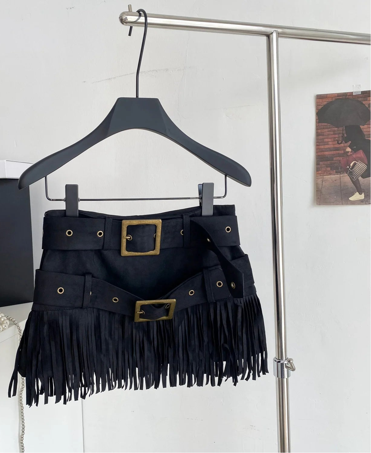 Black fringed belt with gold buckles on a hanger against a white wall.