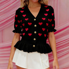 Black cardigan with red heart patterns and white shorts against a pink background