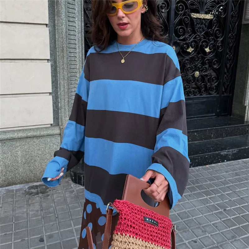 Person wearing a blue and brown striped sweater holding a red and brown bag on a sidewalk.