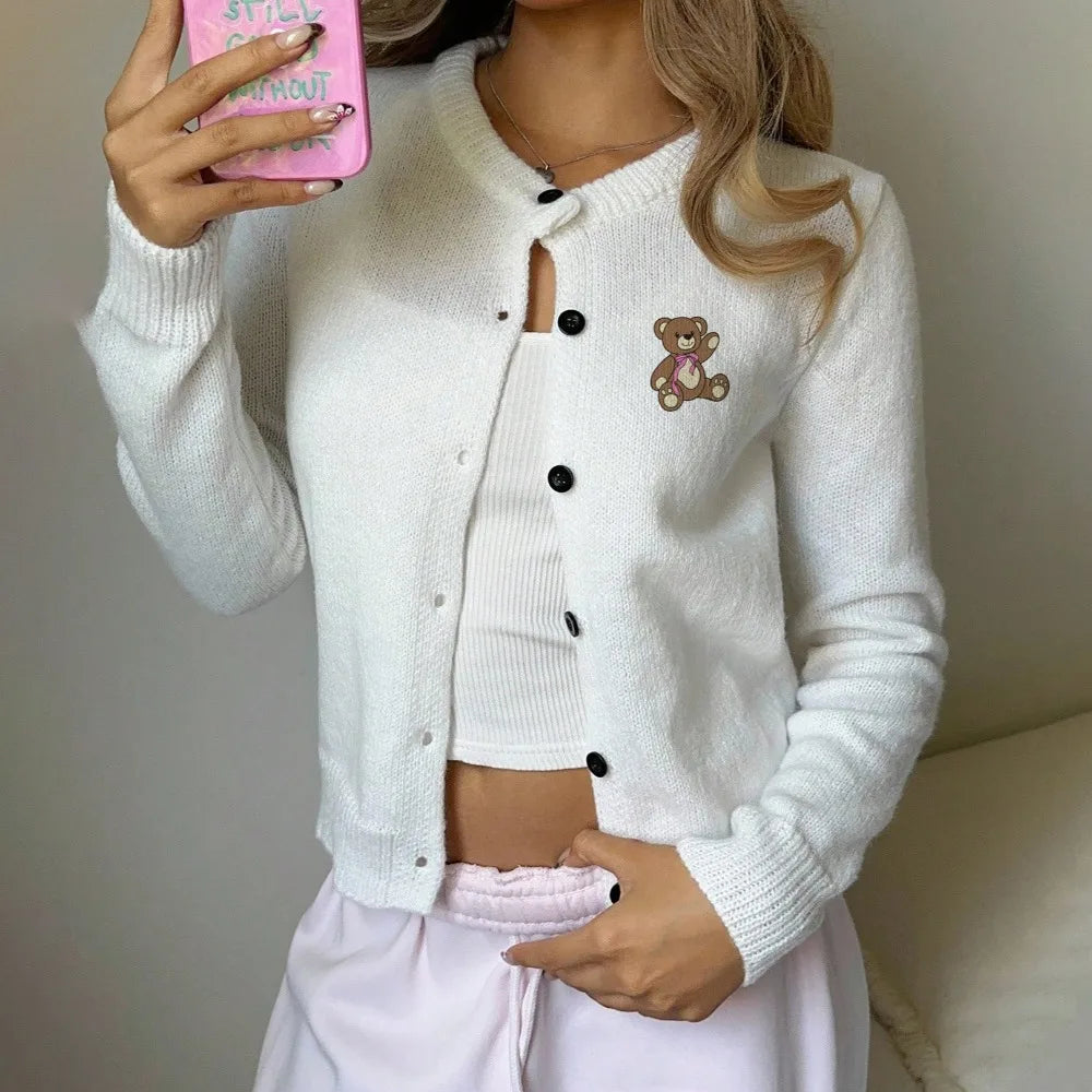 Person wearing a white cardigan with a teddy bear patch, holding a pink phone.