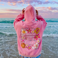 Person wearing a pink hoodie with floral design and text at the beach during sunset.