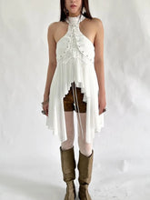 Woman wearing a white halter top dress with sheer panels and brown knee-high boots on a plain background