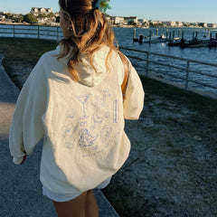 Person wearing a cream-colored hoodie with beach-themed designs by a waterfront.