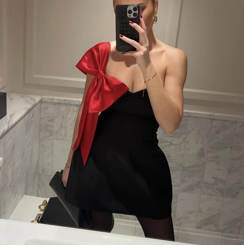 Person wearing a black dress with a large red bow, taking a mirror selfie.