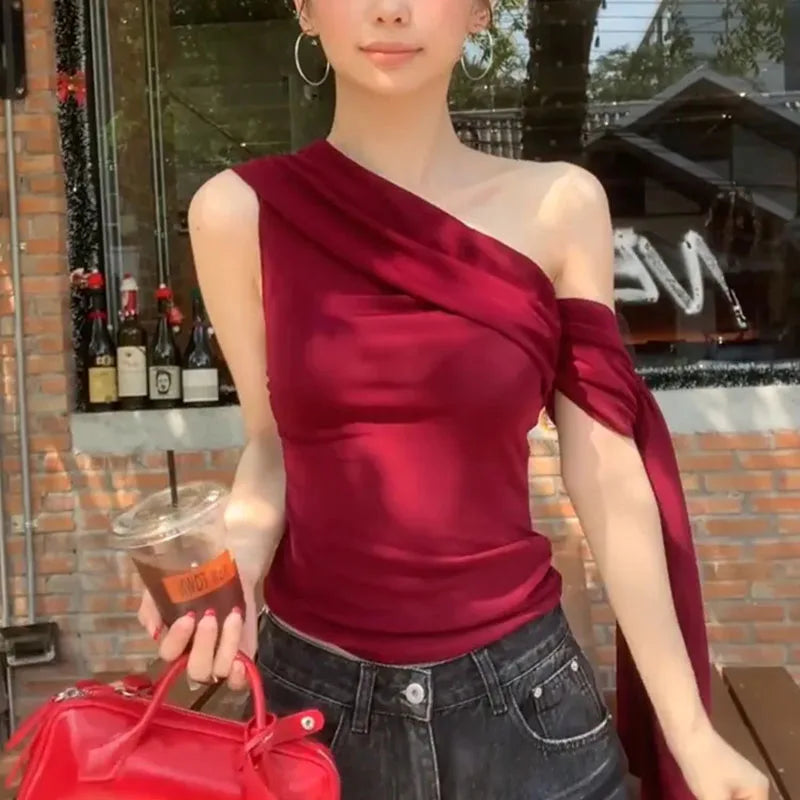 Woman wearing a red one-shoulder top and holding a red handbag and a drink.