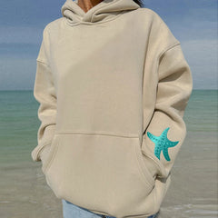 Person wearing a beige hoodie with a turquoise starfish design on a beach