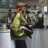 Woman in a green jacket and cap holding a drink in a coffee shop.
