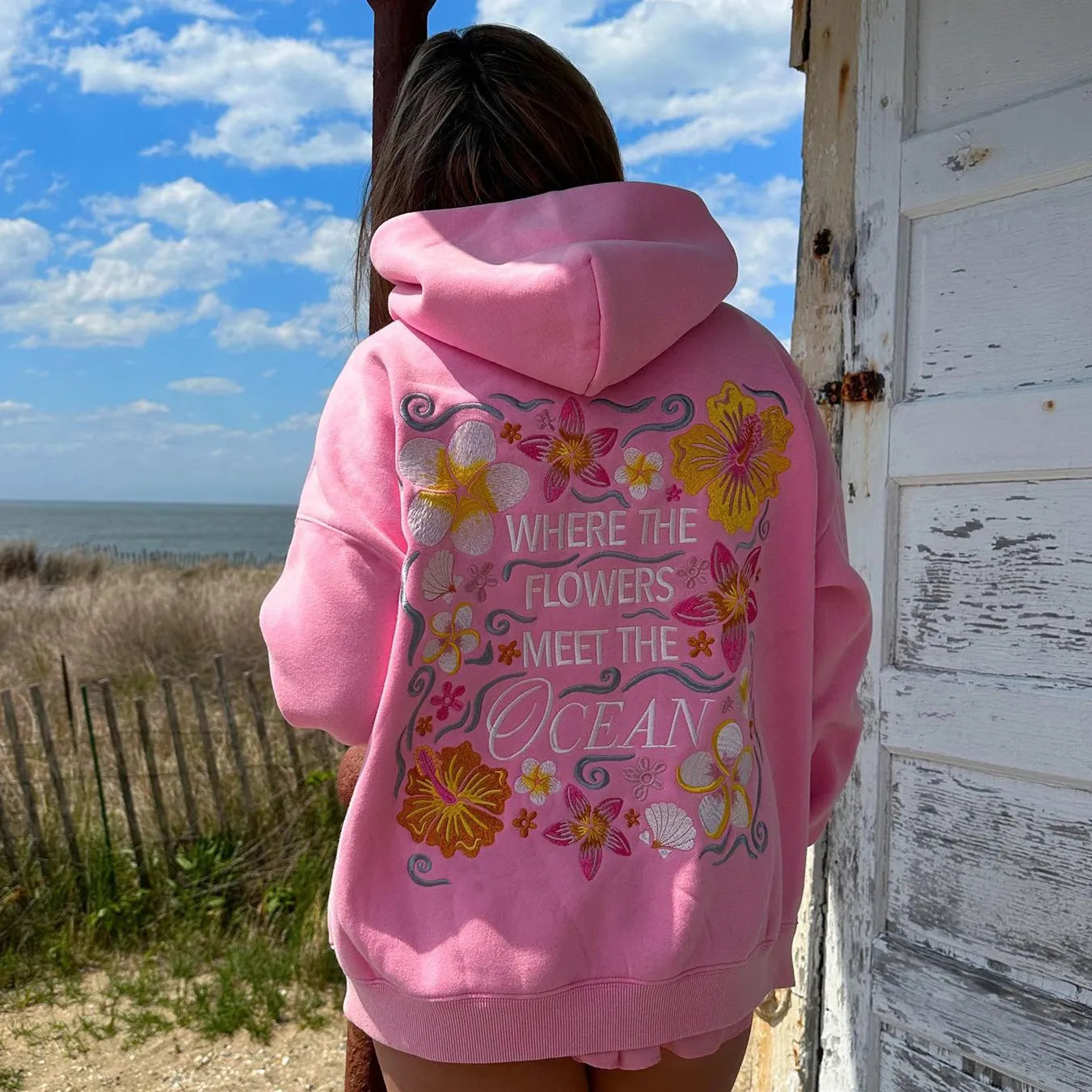 Person wearing a pink hoodie with floral designs and text by a beach.