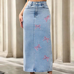 Blue denim skirt with pink bow embroidery worn by a person, standing against a neutral background.