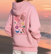 Person wearing a pink hoodie with embroidered designs on the back, standing on a beach at sunset.