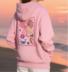 Person wearing a pink hoodie with embroidered designs on the back, standing on a beach at sunset.