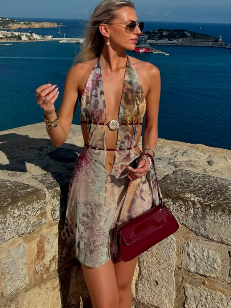 Woman in a floral dress with a red handbag standing on a stone wall by the sea.