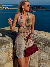 Woman in a floral dress with a red handbag standing on a stone wall by the sea.
