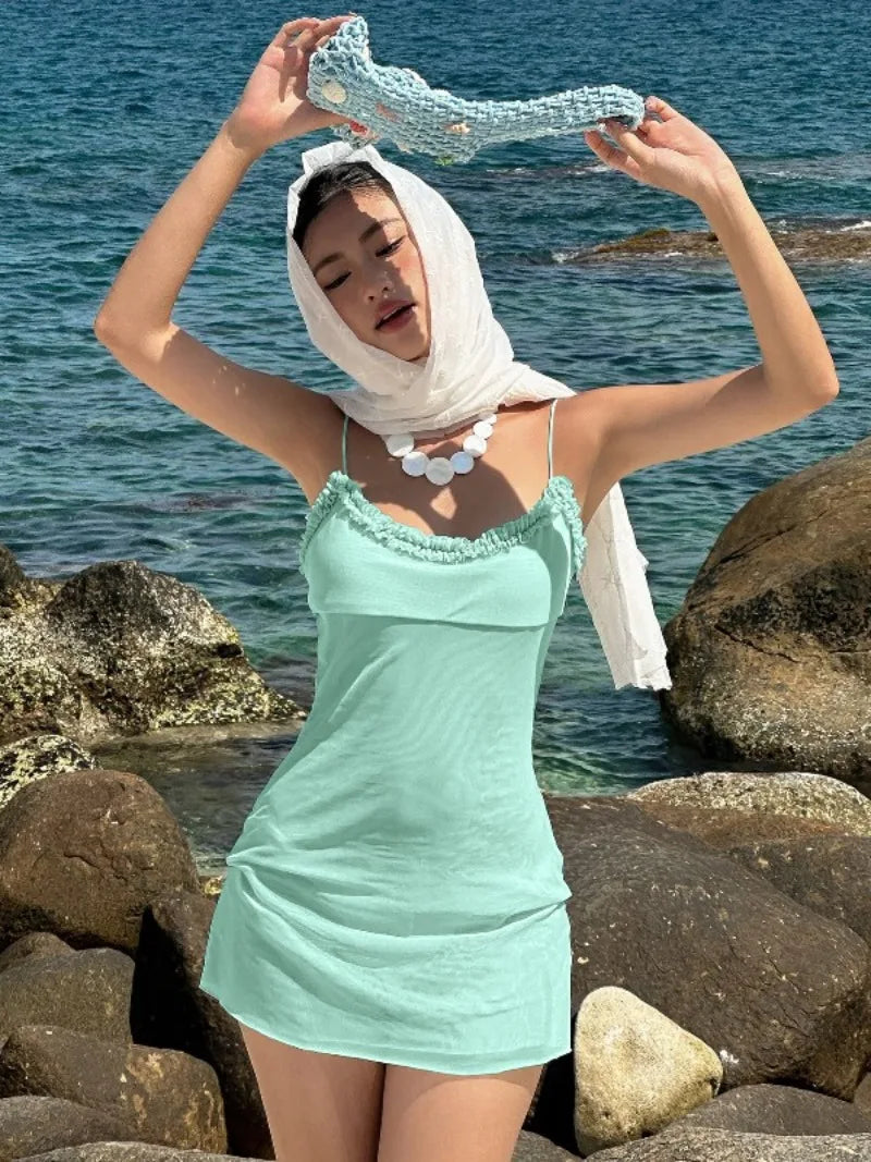 Woman in a light blue dress holding a white headscarf on a rocky beach with ocean in the background