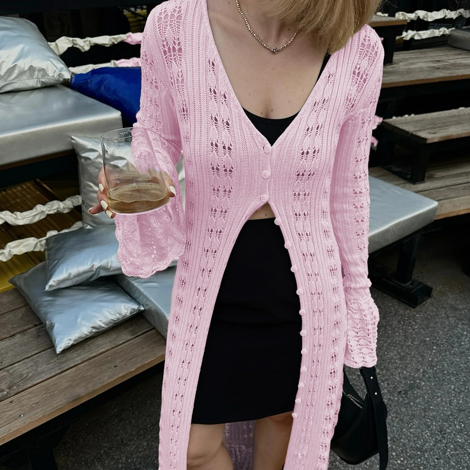 Person wearing a pink cardigan over a black top, holding a drink, with a blurred background.