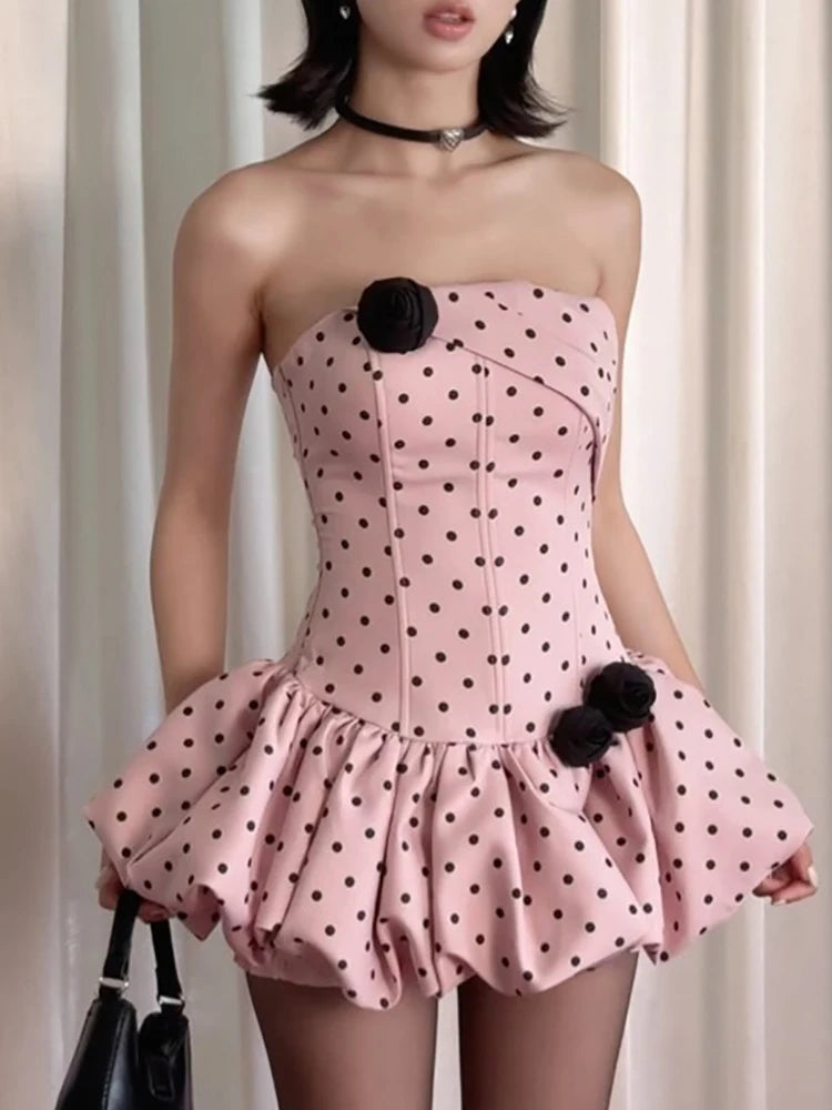 Pink strapless dress with black polka dots and pom-poms worn by a model against a neutral background