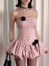 Pink strapless dress with black polka dots and pom-poms worn by a model against a neutral background