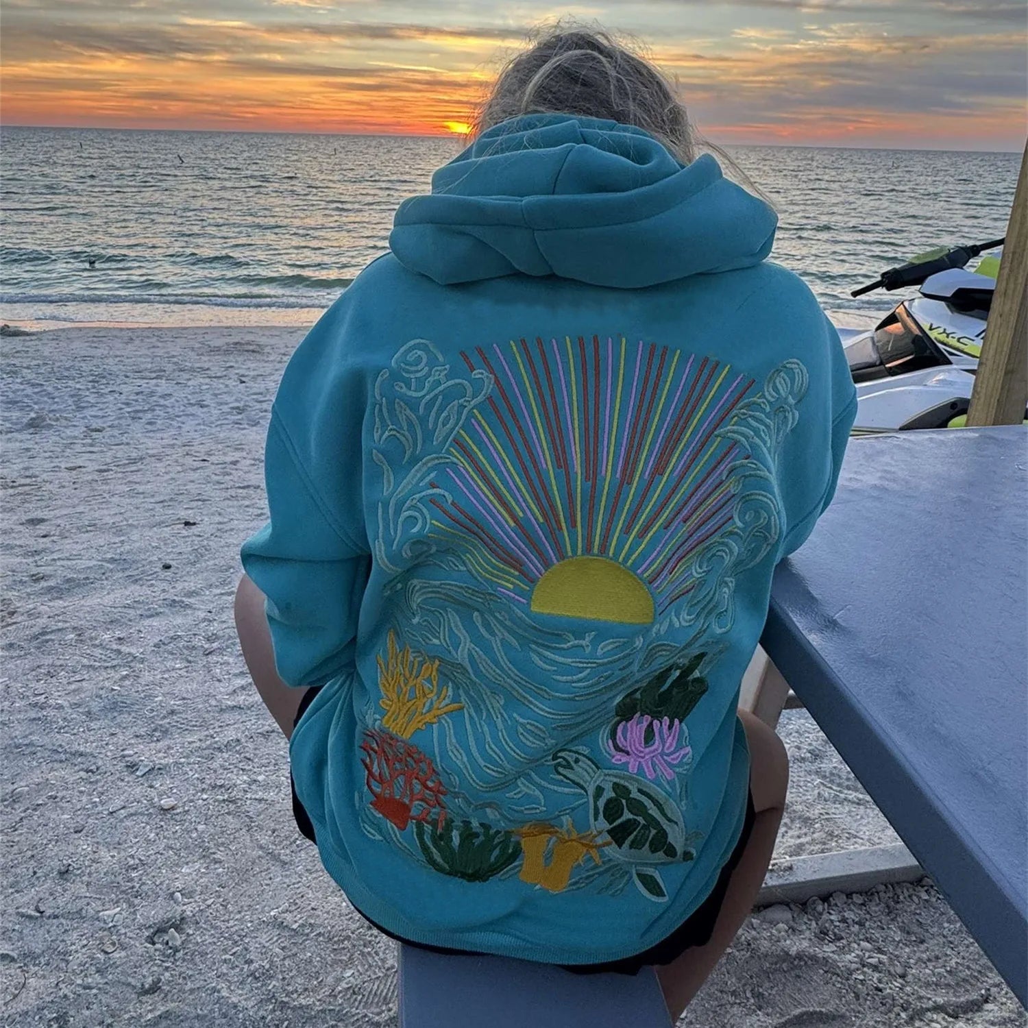 Person wearing a blue hoodie with a colorful design on the beach at sunset