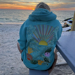 Person wearing a blue hoodie with a colorful design on the beach at sunset