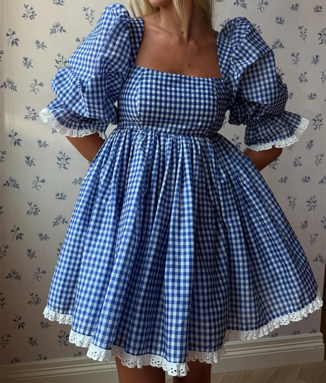Blue and white checkered dress with puffed sleeves and lace trim against a floral wallpaper background.