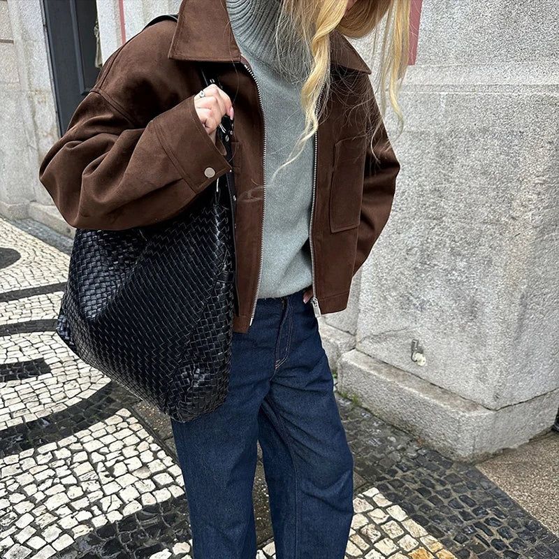 Person wearing a brown jacket and blue jeans, holding a textured black bag on a cobblestone street.