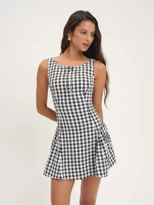 A woman modeling a sleeveless black and white gingham mini dress with a tie-neck detail, set against a plain light background.