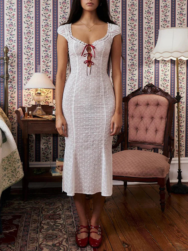 Woman in a white dress with red embroidery standing in a vintage-style room.