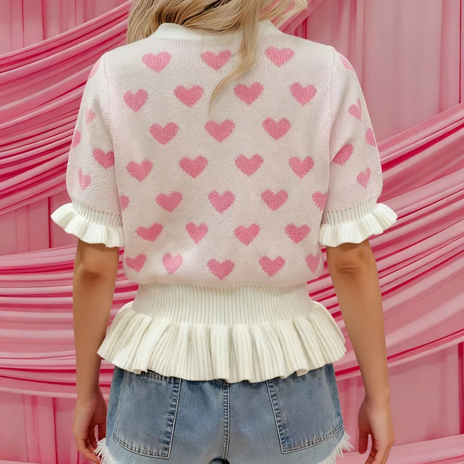 Person wearing a pink sweater with heart patterns and denim shorts against a pink background