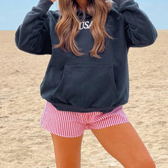 Person wearing a navy sweatshirt with 'USA' text and red and white checkered shorts on a sandy background.