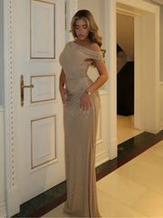 A woman models a floor-length, off-the-shoulder gold sequined gown in an elegant indoor hallway with white paneled doors.