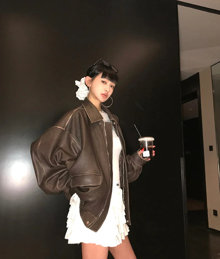 Person wearing a brown leather jacket and white dress holding a cup against a dark wall.