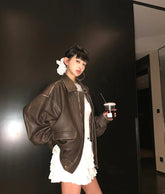 Person wearing a brown leather jacket and white dress holding a cup against a dark wall.