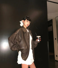 Person wearing a brown leather jacket and white dress holding a cup against a dark wall.