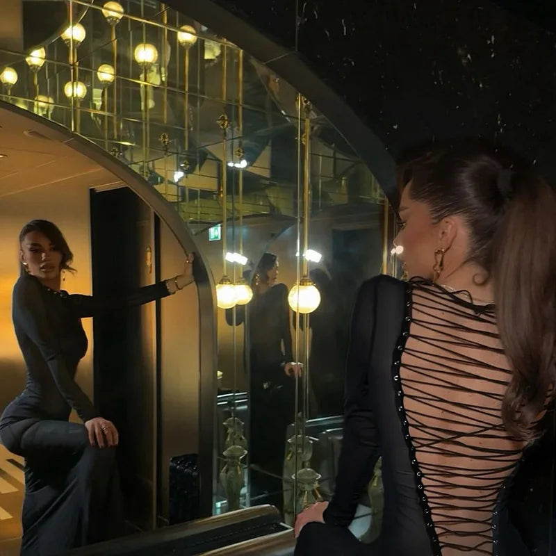 Woman in a black dress with sheer back standing in front of a mirror in a dimly lit room.