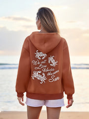 Person wearing a brown hoodie with a quote on the back, standing on a beach.