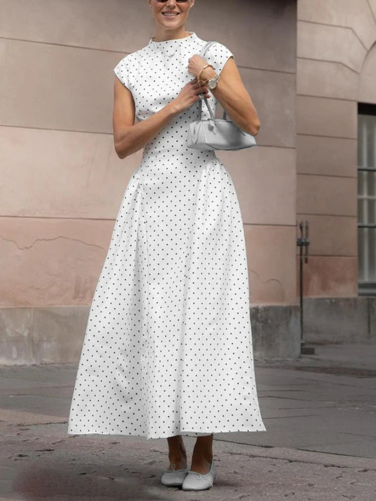 Woman in a white polka dot dress standing against a beige wall.