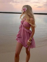 Woman in a pink dress standing on a beach with a flower in her hair at sunset.