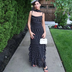 Woman in a black polka dot dress standing on a sidewalk with greenery around.