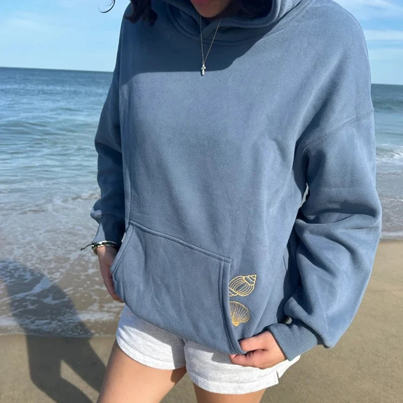Person wearing a blue hoodie with a shell design on a beach