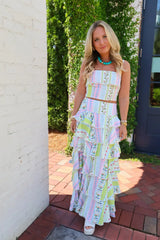 A woman stands outdoors wearing a pastel multicolor tiered maxi dress with a ruffled strapless top and a long layered skirt. The dress has a geometric pattern in pink, blue, green, and white.