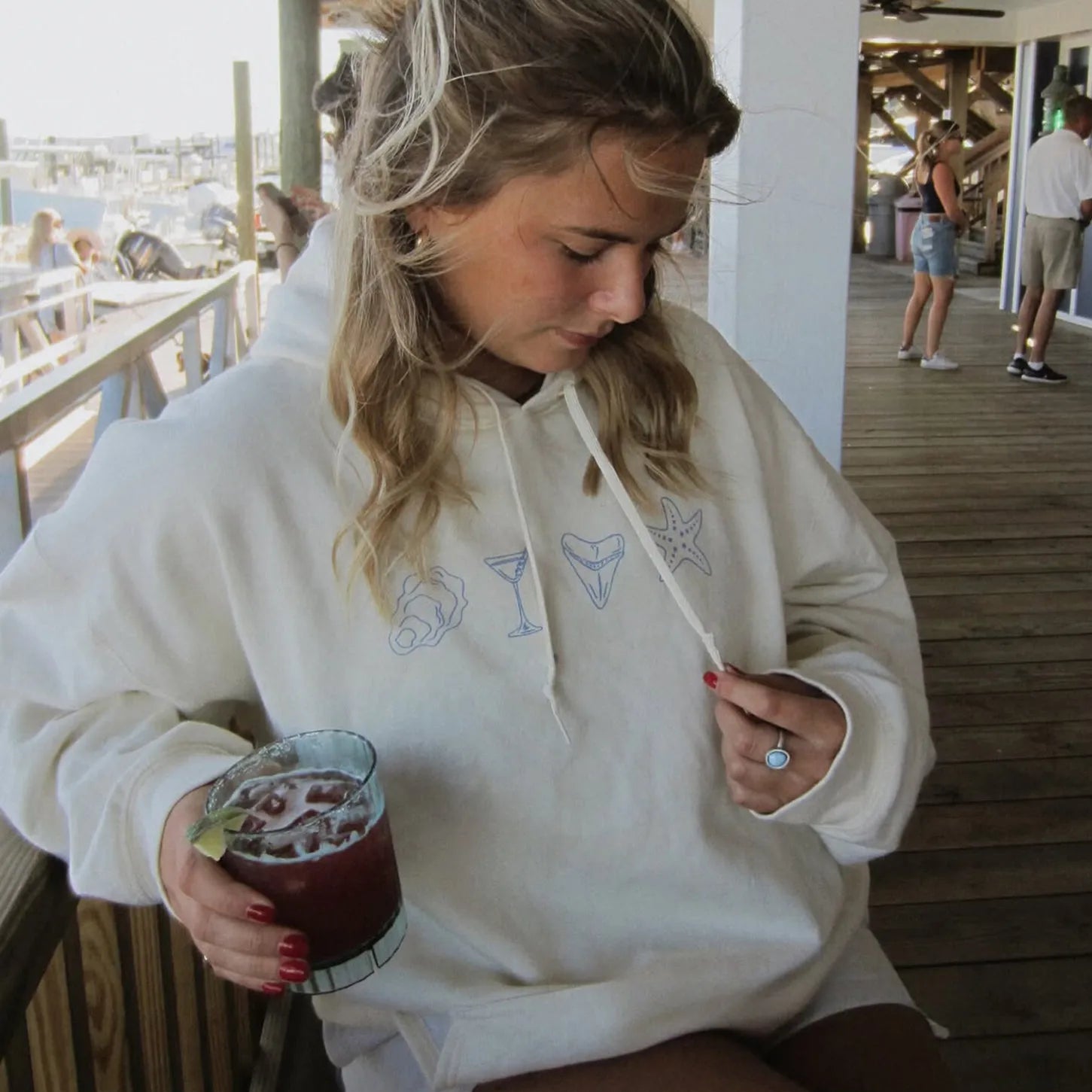 Woman in a white hoodie with embroidered designs, holding a drink on a wooden deck.