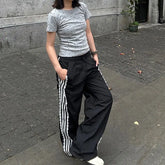 Person wearing a gray t-shirt with white patterns and black pants with white stripes on a street.