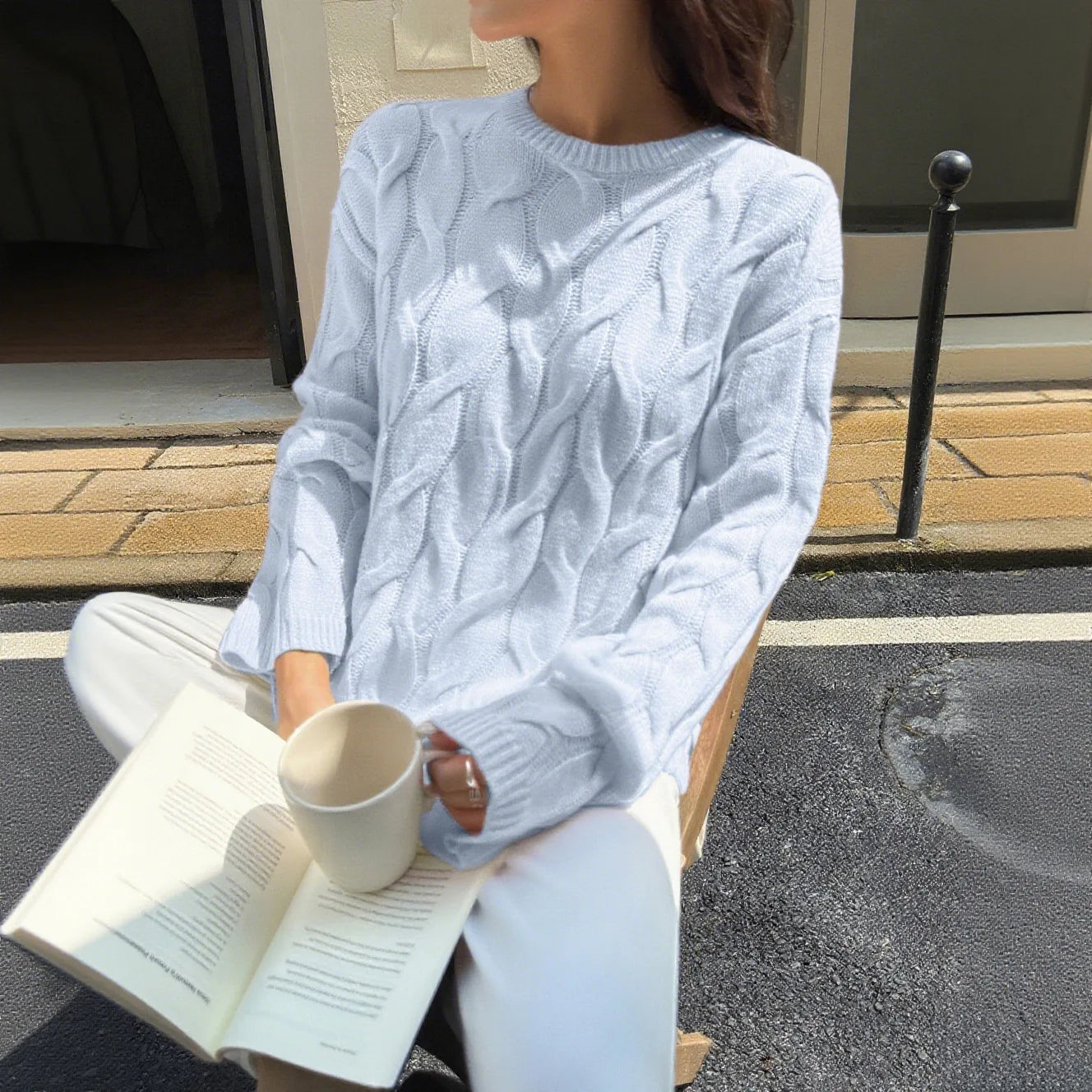 Person wearing a light blue cable knit sweater holding a mug and reading a book outdoors.