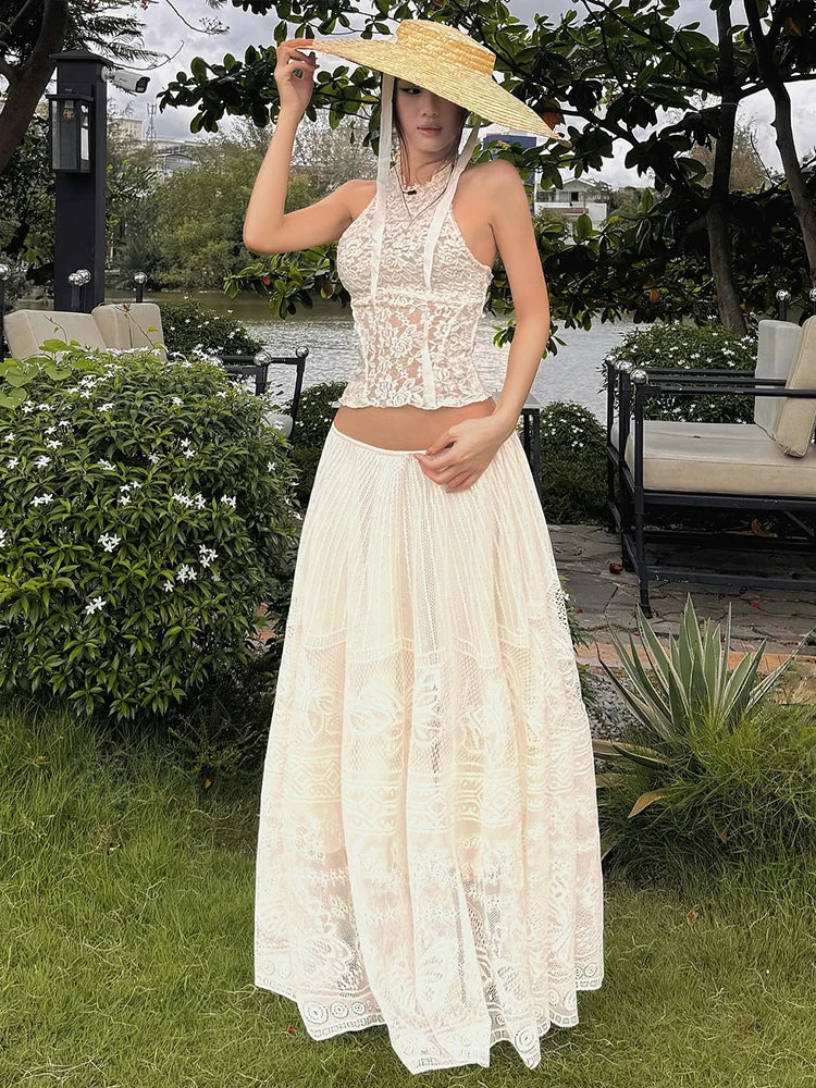 Woman in a lace dress and sun hat standing outdoors near a body of water.