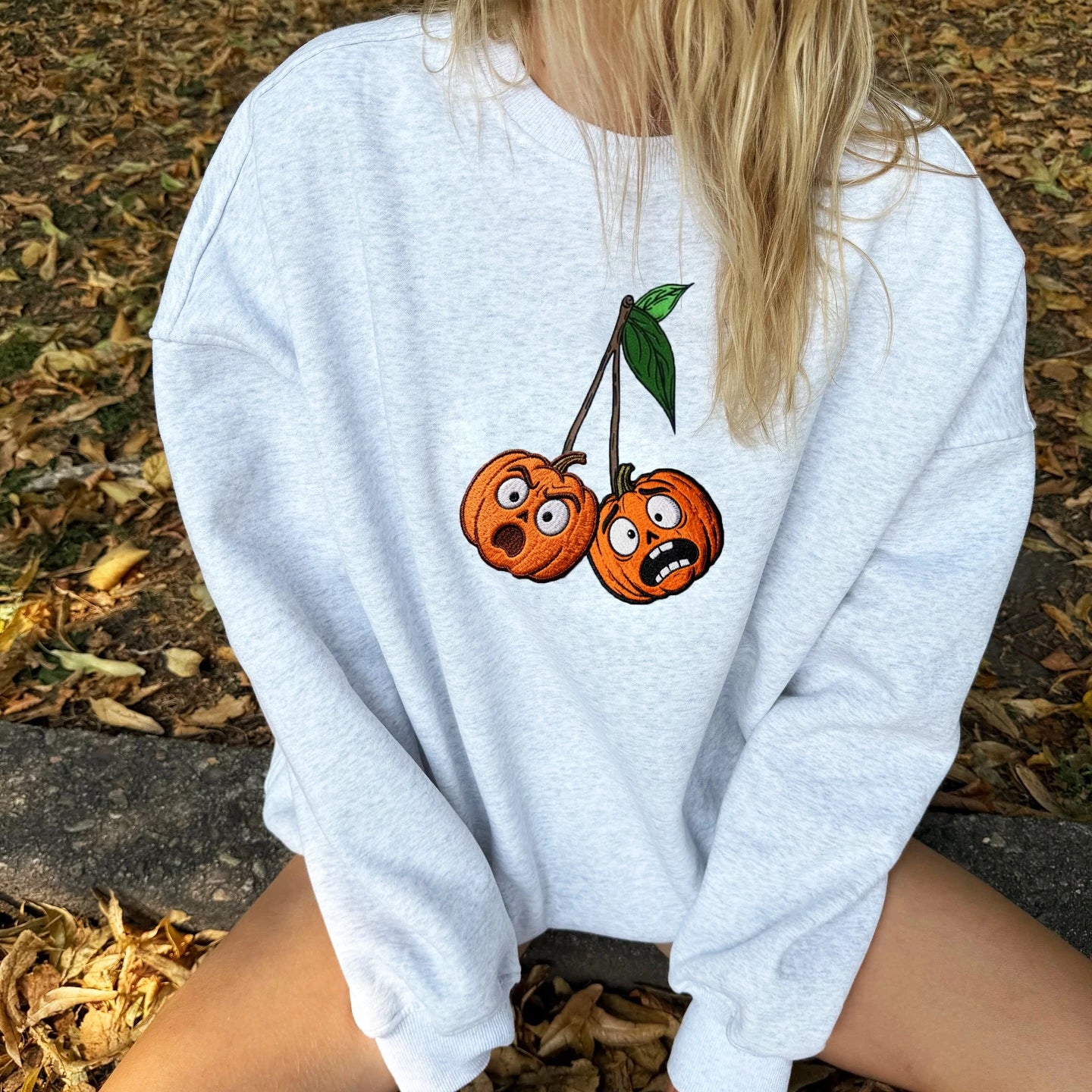 Person wearing a light blue sweatshirt with cartoon pumpkins on the back, sitting on a leaf-covered ground.