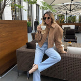 Woman sitting on a outdoor sofa holding a cup, wearing sunglasses and a tan jacket.