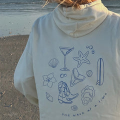 Person wearing a light-colored hoodie with blue beach-themed illustrations on a sandy background